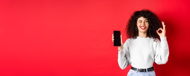 Donna attraente con lo smartphone che mostra il segno giusto e lo schermo del telefono vuoto che consiglia l'app per lo shopping s