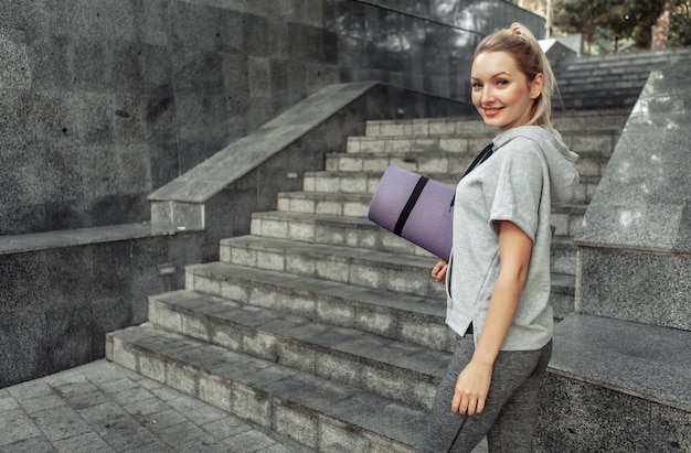 Donna attraente di forma fisica del ritratto con la stuoia arrotolata nelle sue mani all'aperto. Uno stile di vita sano