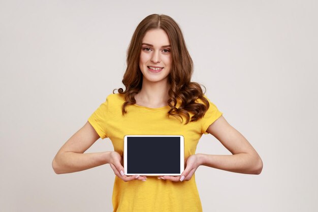 Donna attraente di giovane età che tiene e mostra tablet con schermo vuoto con spazio per la copia del viso sorridente e soddisfatto a trentadue denti per la pubblicità Colpo in studio al coperto isolato su sfondo grigio