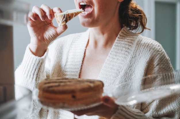 Donna attraente di mezza età in un cardigan accogliente che mangia la torta dal frigorifero in cucina a casa