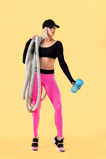 Donna attraente in abiti sportivi rosa e neri con pesanti corde sulle spalle e tenendo agitatore sul muro giallo. Forza e motivazione. Donna sportiva che lavora con corde pesanti.