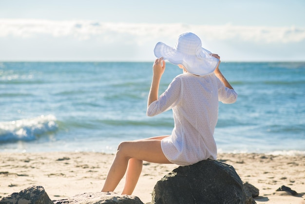 Donna attraente in abito bianco, grande cappello e occhiali che si rilassano sulla spiaggia in una giornata di sole