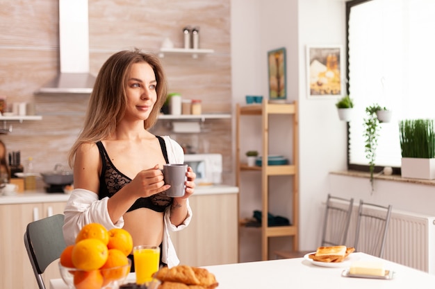 Donna attraente in biancheria intima durante la colazione nella cucina di casa dopo il risveglio.