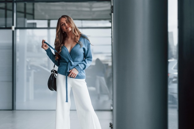 Donna attraente in camicia blu e pantaloni bianchi al chiuso che posano per una macchina fotografica In aeroporto o negozio di auto