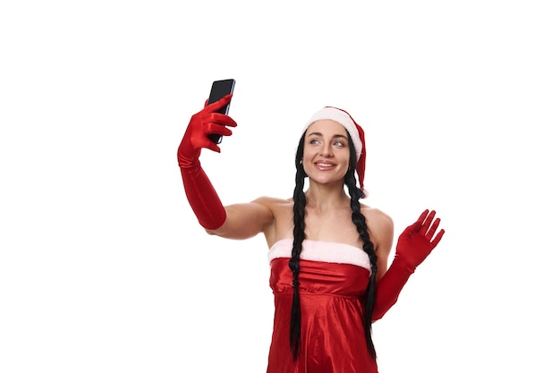 Donna attraente in costume di carnevale di Babbo Natale e tenendo uno smartphone in mano tesa, parlando al telefono, sorridente, videochiamata, facendo selfie, isolato su sfondo bianco. Copia spazio pubblicitario