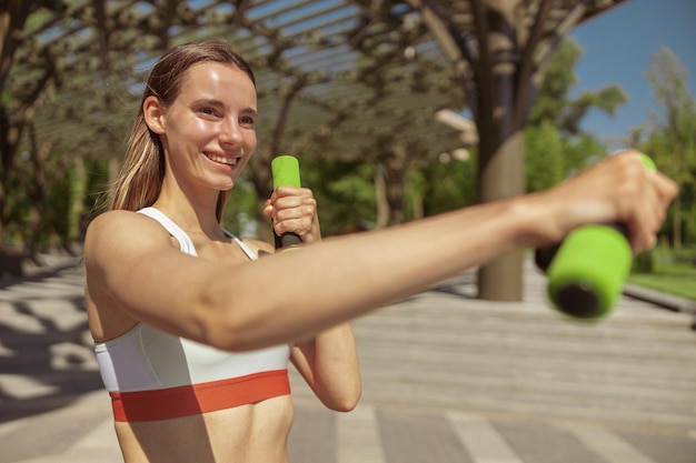 Donna attraente in top sportivo fa esercizi per le braccia con manubri nel parco cittadino