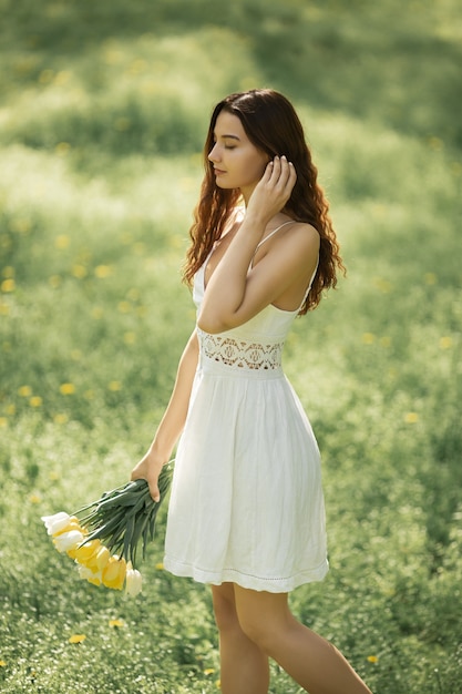 Donna attraente in vestito bianco con un mazzo di fiori primaverili che cammina contro il bokeh della natura