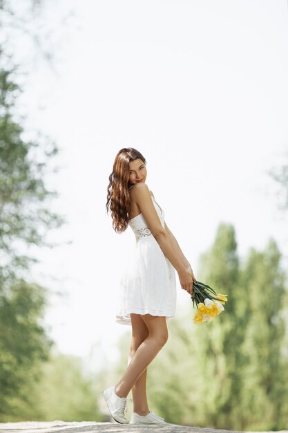 Donna attraente in vestito bianco con un mazzo di fiori primaverili in piedi contro il bokeh della natura