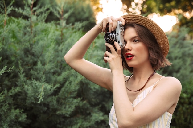 Donna attraente in vestito e cappello di paglia che fa foto sulla retro macchina fotografica all'aperto