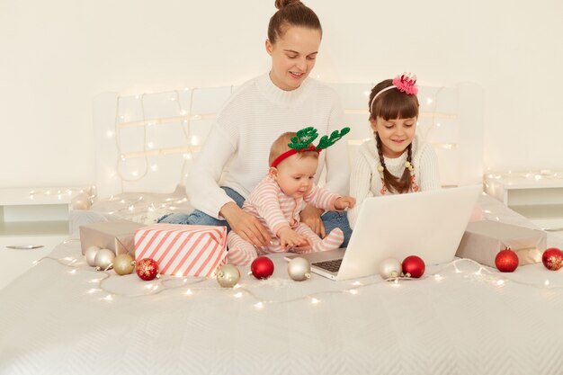 Donna attraente positiva e due bambini che usano il computer portatile per parlare tramite videochiamata mentre sono seduti a letto, guardando il display del notebook, congratulandosi con i loro parenti con le vacanze invernali.