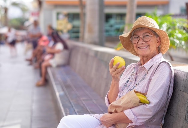 Donna attraente senior allegra che si siede all'aperto nella via della città con un sacchetto di carta con mele e banane che tiene un concetto di mela gialla di pensionato rilassato e mangiare sano