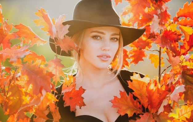 Donna autunnale con foglie d'acero gialle su sfondo autunnale dorato Primo piano ritratto di una bella ragazza con cappello vicino a foglie autunnali Modello femminile autunnale abbastanza romantico