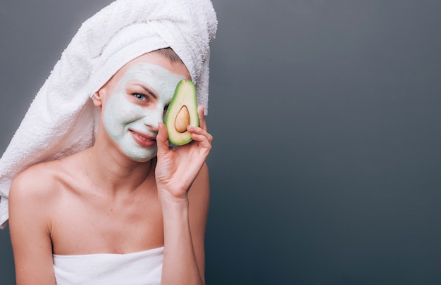 donna avvolta in un asciugamano con una maschera cosmetica sul viso e avocado nelle sue mani