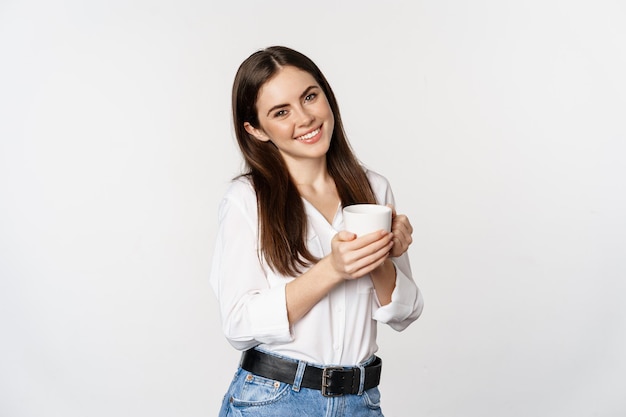 Donna aziendale in piedi con una tazza di tè e caffè e sorridente che beve dalla tazza in piedi felice contro wh...