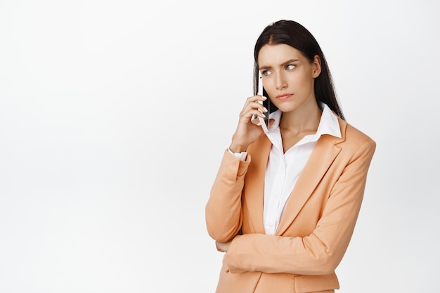 Donna aziendale seria che parla al telefono cellulare accigliata e ascolta con la faccia preoccupata in piedi in tuta su sfondo bianco