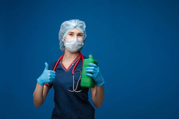 Donna bella in abiti medici bianchi, maschera, guanti blu e phonendoscope sulle spalle con antisettico in grande bottiglia verde, foto isolato su sfondo blu