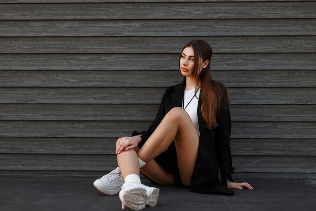 Donna bellissima modella sexy in cappotto nero con scarpe alla moda che si siede vicino a una parete di legno