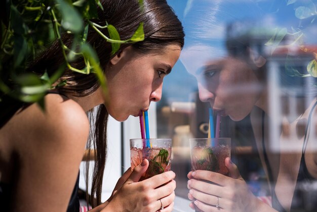 Donna beve bevanda fresca e fredda con la paglia vicino alla finestra signora sexy beve cocktail guardando fuori dalla finestra