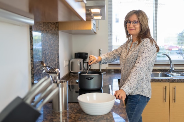 Donna bianca matura che cucina a casa e guarda la telecamera con un atteggiamento felice e sorridente