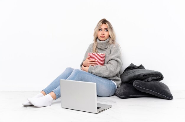 Donna bionda adolescente che mangia popcorn mentre si guarda un film sul computer portatile facendo dubbi gesto mentre si sollevano le spalle