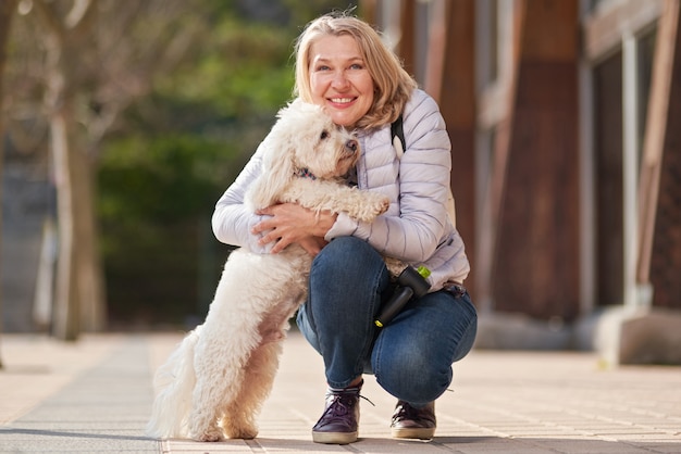 Donna bionda adulta che cammina con il cane bianco lanuginoso nella città di estate.