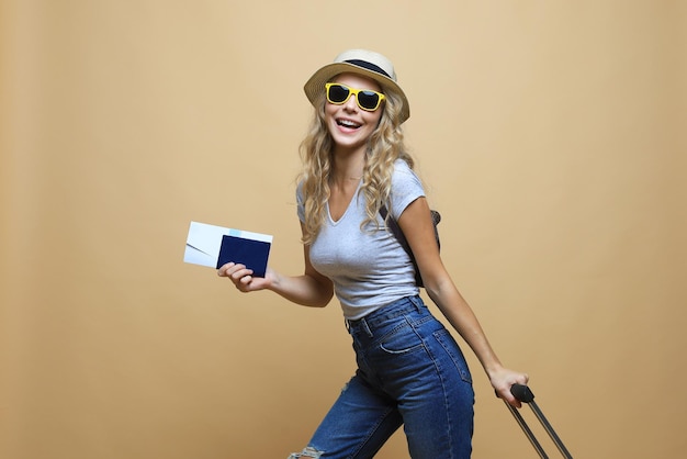 Donna bionda allegra in occhiali da sole in posa con bagagli e in possesso di passaporto con biglietti su sfondo beige.