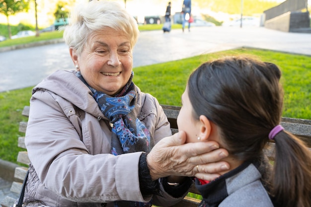 Donna bionda anziana che accarezza la nipote seduta su una panchina nel parco Amore della nonna e della bambina