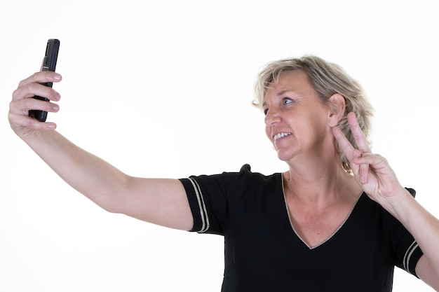 Donna bionda anziana su sfondo bianco che fa selfie al telefono con lo smartphone della fotocamera in mano