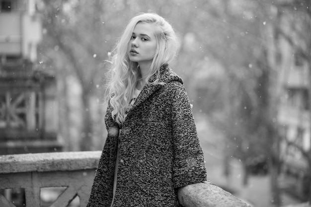 Donna bionda caucasica con capelli ricci, seduta da sola su un balcone. Stagione invernale. Immagine in bianco e nero.