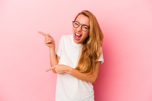 Donna bionda caucasica isolata su sfondo rosa che indica con l'indice uno spazio di copia, esprimendo eccitazione e desiderio.