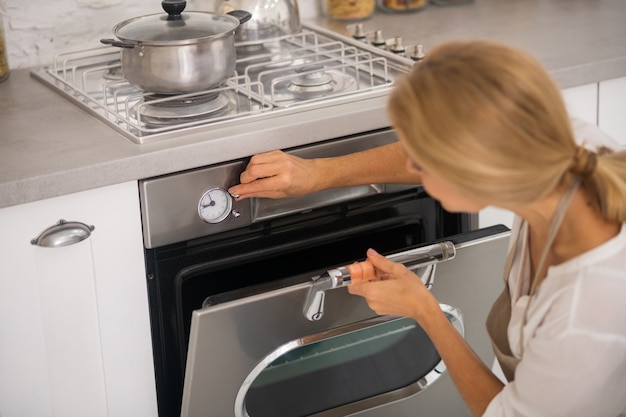 Donna bionda che apre il forno in cucina durante la cottura della cena