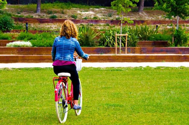 Donna bionda che cammina nel parco con un denim blu su una bicicletta rossa.