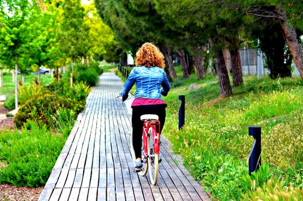 Donna bionda che cammina nel parco con un denim blu su una bicicletta rossa.