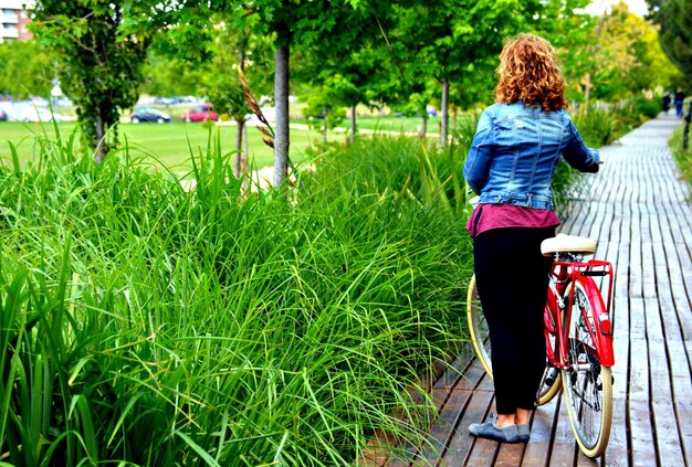 Donna bionda che cammina nel parco con un denim blu su una bicicletta rossa.