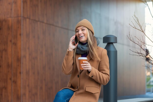 Donna bionda che chatta navigando in internet online sullo smartphone durante la pausa caffè mentre ti rilassi sulla panchina del parco Bella giovane donna carina che utilizza il telefono cellulare e tiene una tazza di caffè