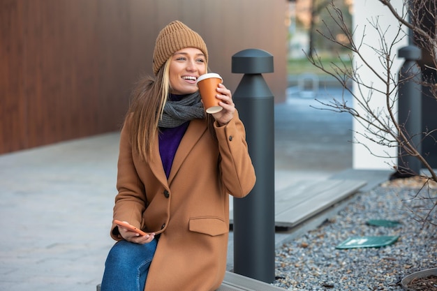 Donna bionda che chatta navigando in internet online sullo smartphone durante la pausa caffè mentre ti rilassi sulla panchina del parco Bella giovane donna carina che utilizza il telefono cellulare e tiene una tazza di caffè