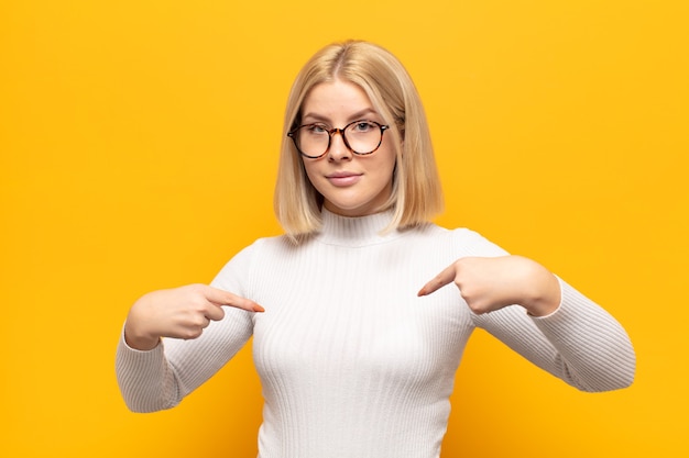 Donna bionda che guarda orgoglioso, positivo e casual che punta al petto con entrambe le mani