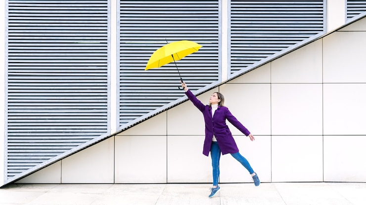 Donna bionda che indossa un cappotto viola che salta con un ombrello giallo su uno sfondo geometrico bianco.