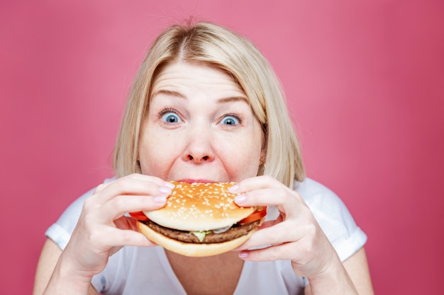 Donna bionda che mangia un grande hamburger succoso. Cibo spazzatura e stile di vita malsano. Sfondo rosa. Avvicinamento.