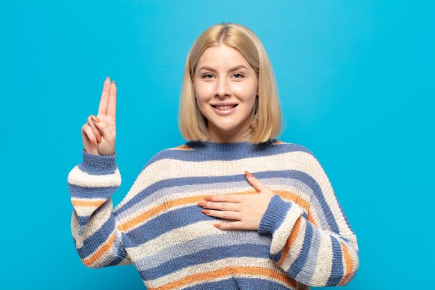 Donna bionda che sembra felice, fiduciosa e degna di fiducia, sorridente e mostrando segno di vittoria, con un atteggiamento positivo