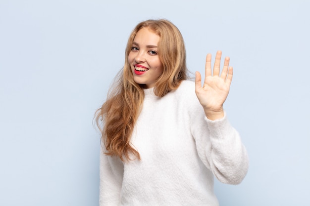 Donna bionda che sorride allegramente e allegramente, agitando la mano, dandoti il benvenuto e salutandoti o salutandoti