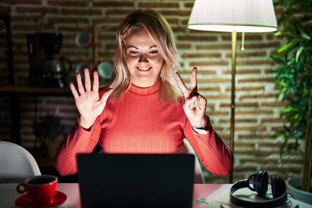 Donna bionda che utilizza il laptop di notte a casa mostrando e indicando con le dita numero sette mentre sorride fiducioso e felice