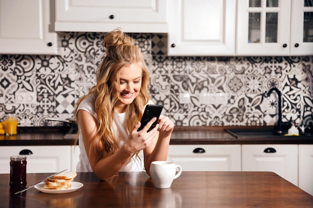 Donna bionda che utilizza smartphone in cucina leggera