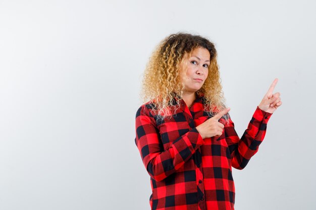 Donna bionda con capelli ricci in camicia a quadri che punta all'angolo in alto a destra e sembra esitante, vista frontale.