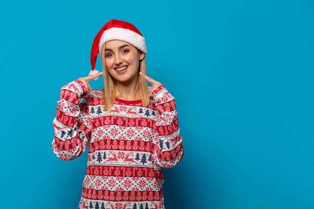 Donna bionda con cappello da Babbo Natale che sorride con sicurezza indicando il proprio ampio sorriso, atteggiamento positivo, rilassato e soddisfatto