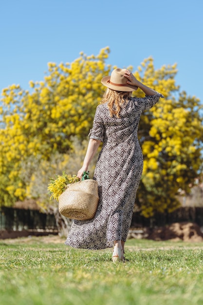 Donna bionda con cappello di paglia e abito lungo in giardino che cammina verso una mimosa con un cesto in mano