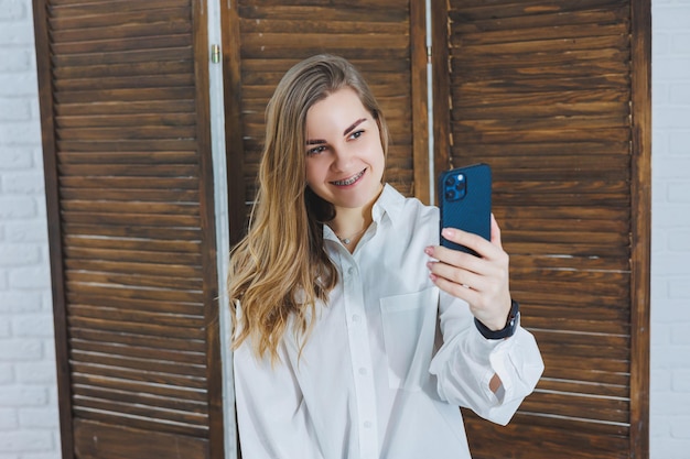 Donna bionda con i capelli lunghi in una camicia bianca e pantaloni neri in posa davanti alla fotocamera del telefono che tiene il telefono e si fa un selfie su uno sfondo isolato