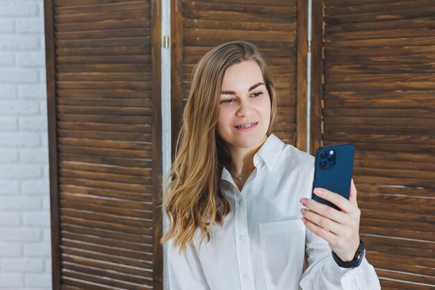 Donna bionda con i capelli lunghi in una camicia bianca e pantaloni neri in posa davanti alla fotocamera del telefono che tiene il telefono e si fa un selfie su uno sfondo isolato