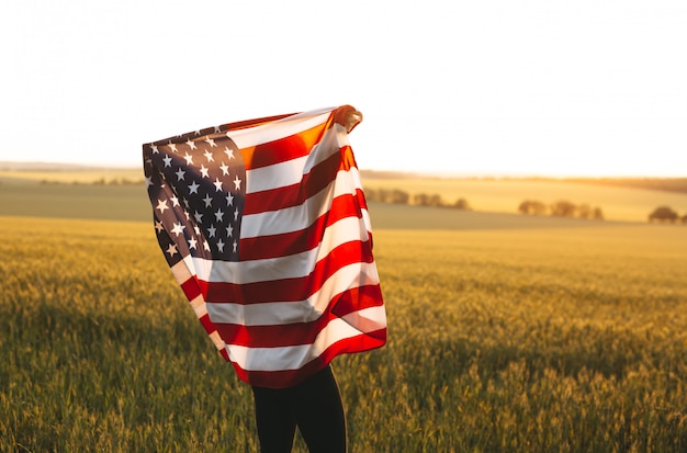 Donna bionda con la bandiera americana in esecuzione in un campo di grano al tramonto. Festa dell'indipendenza, festa patriottica. 4 luglio.
