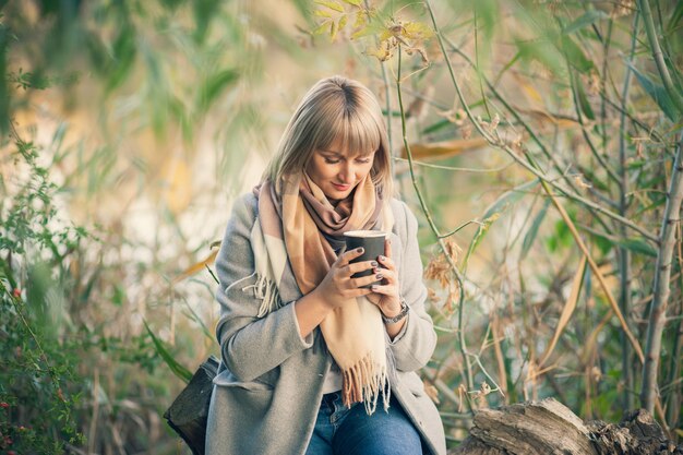 Donna bionda con taglio di capelli corto in cappotto di lana grigia e sciarpa scozzese beve caffè cappuccino con marshmallow. Passeggiate e picnic nella natura nella foresta autunnale